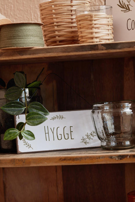 Metalskilt med ordet HYGGE fra Ib Laursen står på en reol med nips