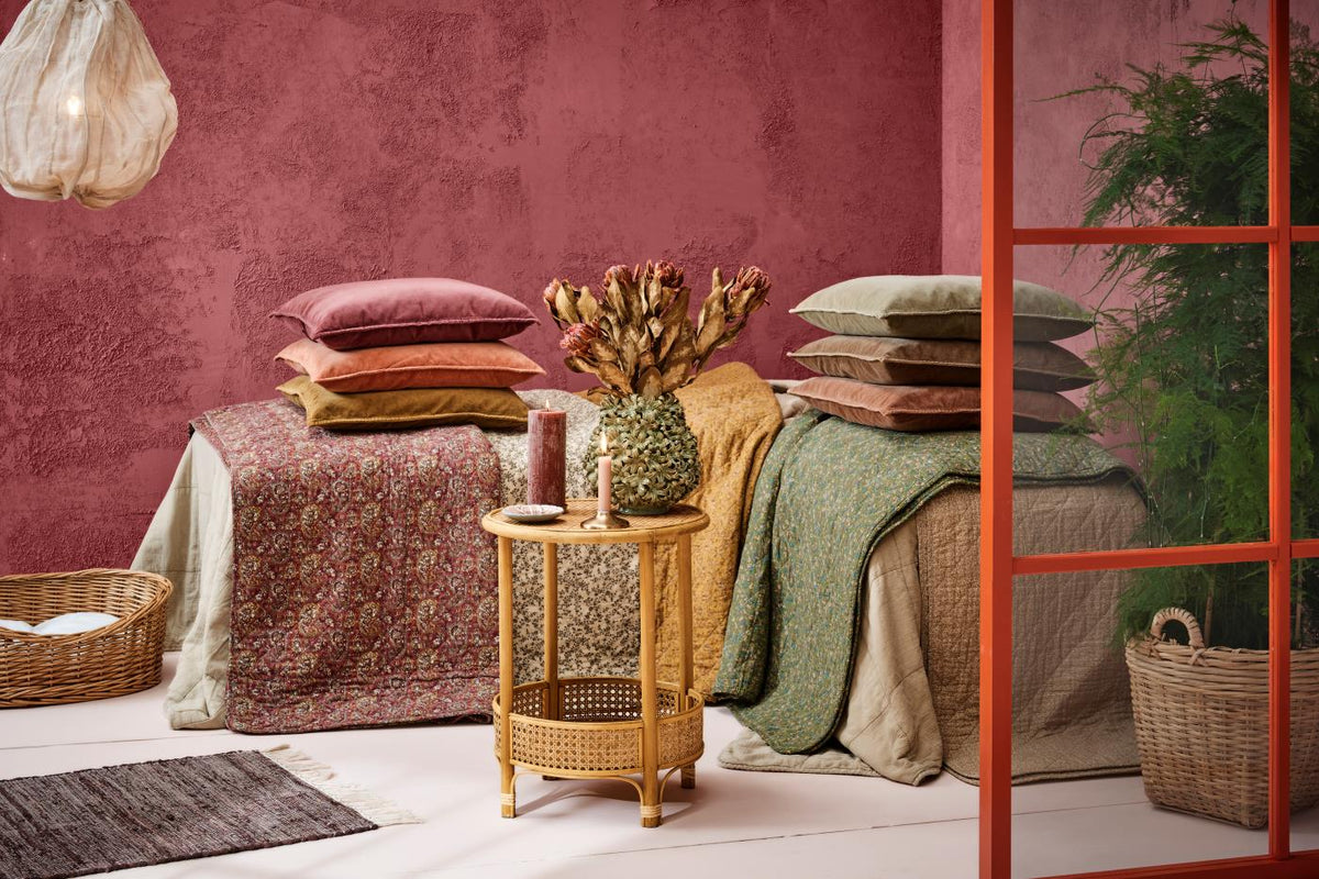 Foldede quilts og puder fra Ib Laursen i forskellige farver og mønstre ved en rosa væg