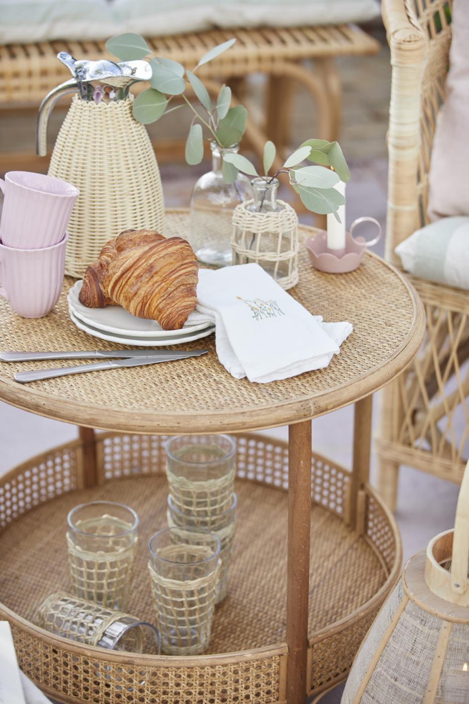 Termokande fra Ib Laursen på rundt fletbord med croissant, glas og blomster i en hyggelig sommerlig borddækning.