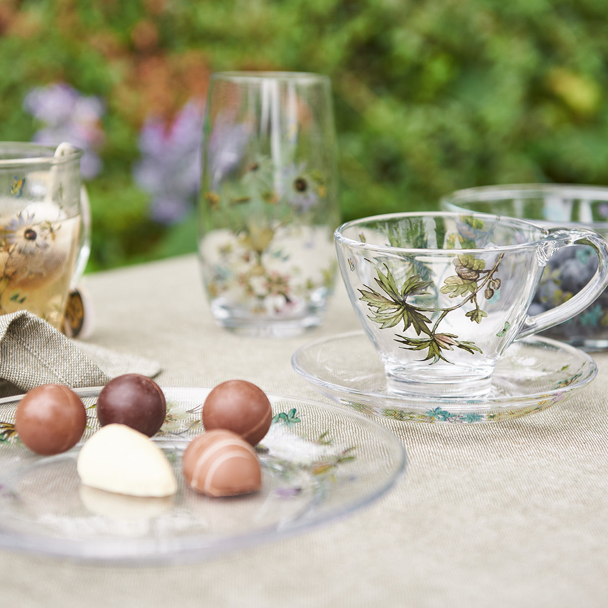 Blomstret glaskop med underkop fra Speedtsberg på sommerligt kaffebord med matchende glas og små chokolader.