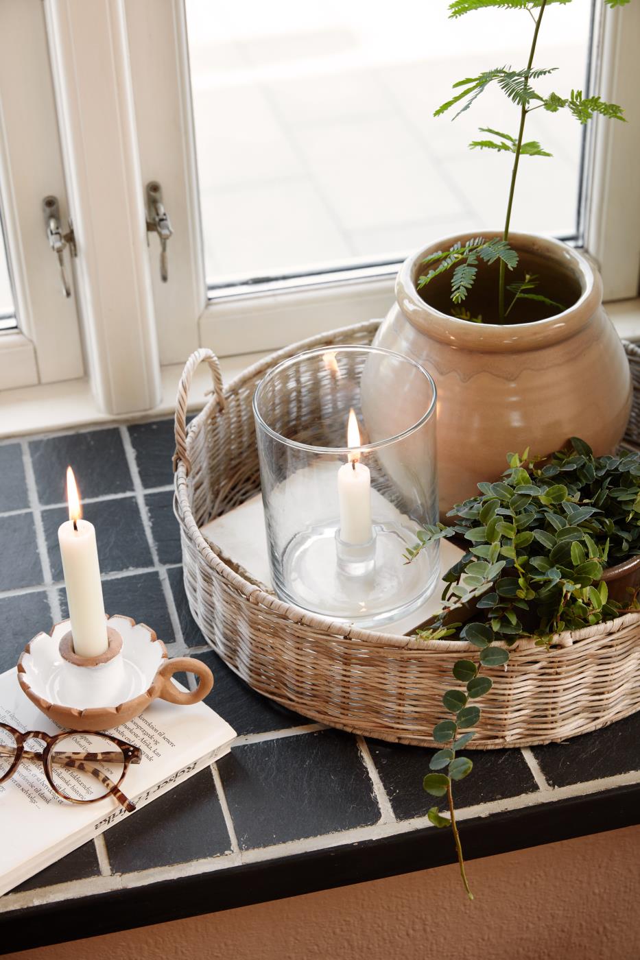 Bred glaslysestage med kronelys på bakke i vindueskarm sammen med grønne planter og naturpynt
